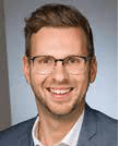 Ein professionelles Porträtfoto eines lächelnden Mannes mit Brille, Anzug und Krawatte.