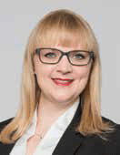 Eine Person mit langen blonden Haaren und Brille, die an einen Lohnsteuerberater erinnert, lächelt in die Kamera. Sie trägt ein helles Hemd und einen dunklen Blazer. Der Hintergrund ist in einem schlichten, hellgrauen Farbton gehalten.