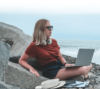 Eine Person in Freizeitkleidung, die an einem Laptop arbeitet, während sie auf felsigem Gelände am Meer sitzt und eine entspannte, aber dennoch konzentrierte Atmosphäre in einer natürlichen HR-Umgebung (Humanressourcen) im Freien ausstrahlt.