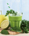 Ein erfrischender grüner Smoothie, garniert mit einem Zweig Petersilie, begleitet von frischen Spinatblättern, einer ganzen Zitrone und einem reifen gelben Apfel, alles vor einem ruhigen blauen Hintergrund.