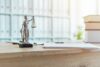 Ein Symbol der Gerechtigkeit: eine Miniaturstatue einer Richterin mit einer Waage, die auf einem Holztisch neben juristischen Dokumenten und Büchern im Hintergrund steht und an die Atmosphäre einer Anwaltskanzlei oder eines Gerichtssaals erinnert.
