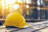 Ein gelber Schutzhelm ruht auf einer Holzoberfläche auf einer in warmes Sonnenlicht getauchten Baustelle.