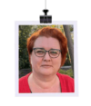Eine Frau mit kurzen roten Haaren und Brille sowie einem roten Oberteil ist im Freien mit Bäumen im Hintergrund abgebildet. Das Bild wird so angezeigt, als ob es oben an einer Halterung befestigt wäre.