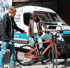An einem sonnigen Tag hilft eine Person einer anderen Person dabei, auf ein rotes Fahrrad neben einem Lieferwagen zu steigen, der offenbar zu einem Bikesharing- oder Fahrradverleihservice gehört.
