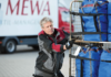 Ein Arbeiter in grau-roter Uniform, Teil des Humanressourcen-Teams, sortiert sorgfältig blaue Taschen auf einem Metallwagen, im Hintergrund ein Lieferwagen.