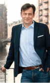 Ein Mann in einem Smart-Casual-Outfit mit blauem Hemd und Blazer steht an einem Geländer mit einer städtischen Uferpromenade im Hintergrund.