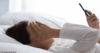 Eine Person, die im Bett liegt, ein Smartphone hält und die andere Hand in einer Geste auf die Stirn legt, die auf Sorgen, Stress oder Kopfschmerzen schließen lässt, möglicherweise besorgt über Probleme im Zusammenhang mit der Personalverwaltung.