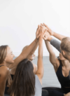 Eine Gruppe von sechs Personen sitzt im Kreis, lächelt und hebt die Hände in einer High-Five-Geste zur Mitte. Sie tragen lässige Trainingskleidung und sitzen auf einem hellen Holzboden vor einem hellen und minimalistischen Hintergrund.