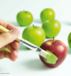Eine Hand, die einen Pinsel hält, trägt einen Strich grüner Farbe auf einen roten Apfel auf und imitiert so die grünen Äpfel im Hintergrund.