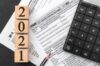 Holzklötze mit der Jahreszahl „2021“, gestapelt auf einem US-amerikanischen Papier. Formular zur individuellen Einkommensteuererklärung für das Jahr 2020, begleitet von einem Bleistift und einem Taschenrechner auf einem Schreibtisch, der Finanzmanagement und Steuererklärung symbolisiert.