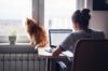 Eine Frau arbeitet an ihrem Laptop neben einem Fenster, ihre Katze sitzt auf der Fensterbank und beide blicken nach draußen.