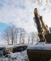 Ein schneebedeckter Baggerarm ragt vor einem Winterhimmel in die Höhe, am Horizont säumen kahle Bäume.