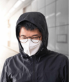 Eine Person mit Brille, weißer Maske und schwarzer Kapuzenjacke, mit geschlossenen Augen und leicht beschlagener Brille.
