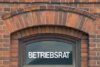 Eine Nahaufnahme eines Fensters mit dem Wort „Betriebsrat“ in weißen Buchstaben auf dem Glas, eingelassen in eine Backsteinmauer mit gewölbter Oberseite.