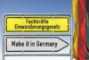 Ein Wegweiser mit zwei Schildern: Auf dem oberen gelben Schild steht auf Deutsch „Fachkräfte Einwanderungsgesetz“ und weist auf den Fachkräftemangel in Deutschland hin, und auf dem unteren weißen Schild steht auf Englisch „Make it in Germany“. Auf der rechten Seite ist vor einem klaren blauen Himmel teilweise die deutsche Flagge zu sehen.