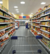 Ein Gang in einem Supermarkt mit Regalen voller verschiedener abgepackter Lebensmittel. Unten im Bild ist teilweise ein Einkaufswagen zu sehen. Der Boden ist gefliest und Leuchtstoffröhren beleuchten den Raum.