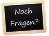 Auf einer Tafel mit Holzrahmen steht mit weißer Kreide der deutsche Satz „Noch Fragen?“, was auf Englisch „Any questions?“ bedeutet.