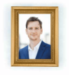 Ein gerahmtes Porträt einer Person mit kurzen Haaren, die einen blauen Anzug und ein weißes Hemd trägt, vor einem unscharfen Hintergrund. Der Rahmen ist golden mit einem strukturierten Rand.