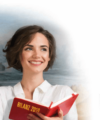 Eine Person mit kurzen braunen Haaren lächelt und hält ein rotes Buch mit dem Titel „BILANZ 2019“. Im Hintergrund ist ein unscharfes Landschaftsgemälde zu sehen.