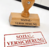 Ein Holzstempel mit dem aufgedruckten Text „SOZIAL-VERSICHERUNG“. Darunter befindet sich in roter Farbe auf weißem Untergrund der Abdruck des Stempels mit dem ebenfalls aufgedruckten Text „SOZIAL-VERSICHERUNG“.