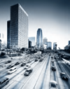 Eine Autobahn mit verschwommener Bewegung und zahlreichen Autos im Vordergrund führt zu einer Stadtlandschaft mit modernen, hohen Wolkenkratzern, darunter markante Hochhäuser. Der monochromatische blaugraue Ton verleiht der Szene ein elegantes, urbanes Flair.