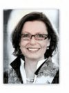 Eine Frau mit kurzen braunen Haaren lächelt in die Kamera. Sie trägt eine Brille, einen dunklen Blazer, ein weißes Hemd mit Kragen und einen Schal mit Paisleymuster. Der Hintergrund ist unscharf.