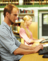 Zwei Personen, ein Mann und eine Frau, sitzen an einem Tisch in einer Bibliothek und arbeiten jeweils an einem Computer. Der Mann im Vordergrund konzentriert sich auf seinen Bildschirm, während die Frau im Hintergrund ebenfalls tippt. Hinter ihnen sind Bücherregale und eine Tür zu sehen.
