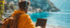 Eine Person in einem gelben Kapuzenpullover und Kopfhörern arbeitet draußen an einem Laptop mit malerischer Aussicht auf einen See und Berge. Ein Rucksack liegt in der Nähe und neben dem See sind unter einem klaren Himmel ein paar Häuser zu sehen.
