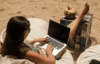 Eine Person entspannt sich auf einem Sitzsack am Strand und tippt auf einem Laptop. Ihre Füße ruhen auf einem verwitterten Holztisch neben ihr. Auf dem Tisch steht ein Kokosnussgetränk mit Strohhalm. Sie trägt einen Badeanzug und eine Sonnenbrille.