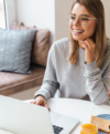 Eine Frau mit Brille und grauem Pullover sitzt lächelnd an einem Tisch und recherchiert fleißig auf ihrem Laptop zum Thema Altersvorsorge. Auf dem Tisch stehen ein Glas Orangensaft und ein aufgeschlagenes Buch, im Hintergrund steht eine gemütliche Couch mit Kissen.
