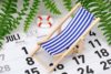 Ein kleiner blau-weiß gestreifter Liegestuhl steht auf einem aufgeklappten Kalender, der bis Juli reicht, und symbolisiert so die Erreichbarkeit im Urlaub. Farnblätter rahmen die Szene ein, im Hintergrund ist ein kleiner Rettungsring zu sehen, der eine heitere Urlaubsstimmung erzeugt.