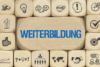 Holzklötze mit verschiedenen Symbolen umgeben das Wort „WEITERBILDUNG“ in Blau. Zu den Symbolen gehören eine Glühbirne, Diagramme, ein Händedruck und eine Weltkarte, die Lernen, Wachstum, Kommunikation und die Anerkennung beruflicher Qualifikationen symbolisieren.
