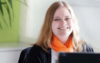 Eine Person mit langen hellen Haaren und einem leuchtend orangefarbenen Schal sitzt lächelnd an einem Schreibtisch und ist konzentriert mit Entgeltabrechnung beschäftigt. Der Hintergrund ist sanft in Grün- und Weißtönen beleuchtet.
