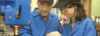 Zwei Personen in blauer Arbeitskleidung und Schutzbrillen arbeiten an einer blauen Industriemaschine. Sie sind konzentriert auf ihre Aufgabe, während eine Person etwas erklärt. Beide tragen schwarze Schutzkappen.