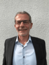 Ein lächelnder Mann mit kurzen grauen Haaren und Brille trägt einen dunklen Blazer über einem hellblauen Hemd. Er steht vor einer strukturierten hellgrauen Wand.