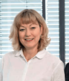 Eine Frau mit schulterlangen blonden Haaren lächelt in die Kamera. Sie trägt eine weiße Bluse und steht vor einem Hintergrund mit vertikalen Jalousien.