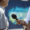 Person, die ein Mikrofon hält und ein Interview mit einer anderen Person führt. Der Hintergrund ist verschwommen und weist Andeutungen von Grün- und Blautönen auf.