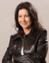 Eine Frau mit langen dunklen Haaren und einer schwarzen Lederjacke lächelt und schaut zur Seite. Sie steht mit verschränkten Armen vor einem schlichten Hintergrund.
