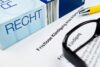 Das Bild zeigt ein Dokument mit dem Titel „Fristlose Kündigung“ auf Deutsch, begleitet von einem blauen Buch mit der Aufschrift „Recht“, einem Stift und einer schwarzen Brille, was auf ein Szenario einer rechtlichen oder arbeitsrechtlichen Kündigung hindeutet, möglicherweise auf „Kündigung aus politischen Gründen“.