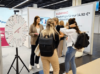 Auf der Zukunft Personal Europe versammeln sich Menschen um ein Spinnrad am Stand, der E-Learning-Dienste für 14,90 Euro anpreist. Eine Frau im Anzug unterhält sich mit drei Frauen, die Rucksäcke tragen, und schafft so eine lockere und doch anregende Atmosphäre.