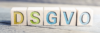 Holzklötze mit der Aufschrift „DS‑GVO“ in bunter Schriftart sind kunstvoll auf einer Holzoberfläche angeordnet, vor einem sanft unscharfen Hintergrund.