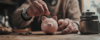 Eine ältere Person wirft eine Münze in ein kleines rosa Sparschwein auf einem Holztisch, vielleicht denkt sie über ihre Lohnsteuerpflicht nach. In der Nähe liegen ein Stapel Münzen und ein Metallgefäß. Die Atmosphäre ist warm und rustikal.