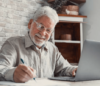 Älterer Mann mit Brille schreibt an einem Schreibtisch auf Papier, lächelt und benutzt einen Laptop. Er sitzt in einem gemütlichen Raum mit einer Ziegelwand im Hintergrund und Regalen voller Körbe.