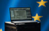 Auf einem Ständer steht ein Laptop, auf dessen Bildschirm eine Programmierschnittstelle angezeigt wird. Im Hintergrund ist eine verschwommene Flagge mit gelben Sternen auf blauem Grund zu sehen, die an die Flagge der Europäischen Union erinnert.