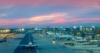 Ein Flughafen in der Abenddämmerung mit mehreren Flugzeugen, die an den Gates parken. Der Himmel ist in rosa und violette Farbtöne getaucht. Die Flughafenlichter sind an und die Landebahn ist sichtbar, was eine lebhafte und geschäftige Atmosphäre schafft.