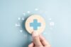 Hand hält ein rundes Holzsymbol mit blauem Kreuz auf hellblauem Hintergrund, umgeben von medizinischen Icons – Symbolbild für private Kranken- und Pflegeversicherung sowie Gesundheitsvorsorge.