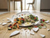 Zerbrochener Teller mit Essen auf Boden im Homeoffice symbolisiert Unfallrisiko bei Mittagspause