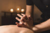 Professionelle Massage-Szene: Eine Therapeutin führt eine Rückenmassage durch. Im Vordergrund sind die Hände der Masseurin auf dem Rücken des Klienten zu sehen. Im Hintergrund brennen drei Kerzen auf einem Tisch und schaffen eine entspannte, ruhige Atmosphäre in einem Behandlungsraum. Empfehlung für WordPress: