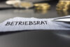 Nahaufnahme eines Schreibtisches mit einem Notizzettel, auf dem handschriftlich "BETRIEBSRAT" steht. Im unscharfen Hintergrund sind gestapelte Euromünzen und ein Taschenrechner zu sehen, was den Bezug zu finanziellen und vergütungsrelevanten Themen symbolisiert.