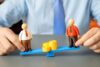 Hände einer Person im Hemd halten eine blaue Wippe mit zwei Spielfiguren; Münzstapel kippen das Gleichgewicht und symbolisieren Gehaltsgefälle sowie ungleiche Bezahlung.